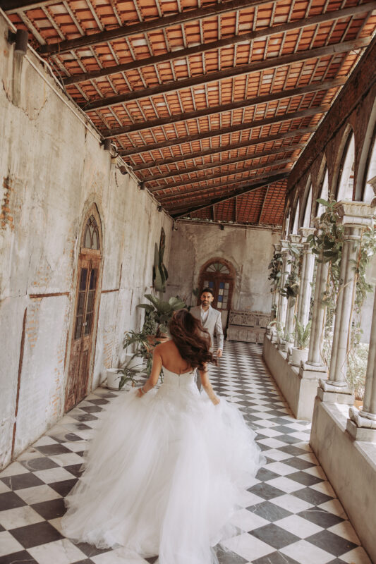 Bride running toward groom in cloister with chequered marble floor, Benedict Studio Bangkok